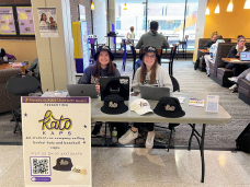 Two students wearing hats sitting at a table with hats on the table and a sign in front of the table that says Kato Kaps