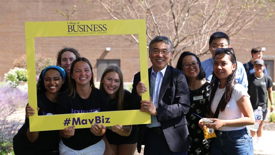 Seung Bach, Dean of the College of Business, posing with students outside on campus holding a yellow picture frame that says, "#MavBiz"