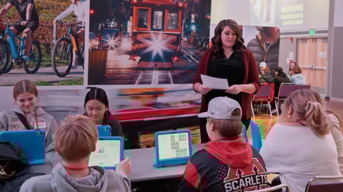 Students sitting down at a table while a volunteer is helping them with the JA's simulation module software at the Junior Achievement North Finance Park Mobile Days held in the CSU Ballroom