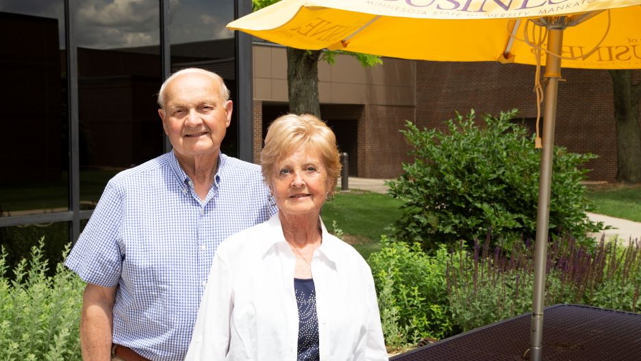 Daryl and Karyl Henze posing outside the College of Business building next to a purple table with a yellow College of Business umbrella