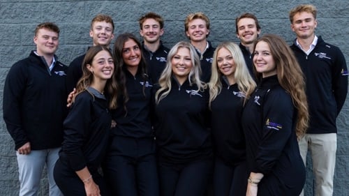 Group of students standing in front of a gray brick wall with matching black jackets.