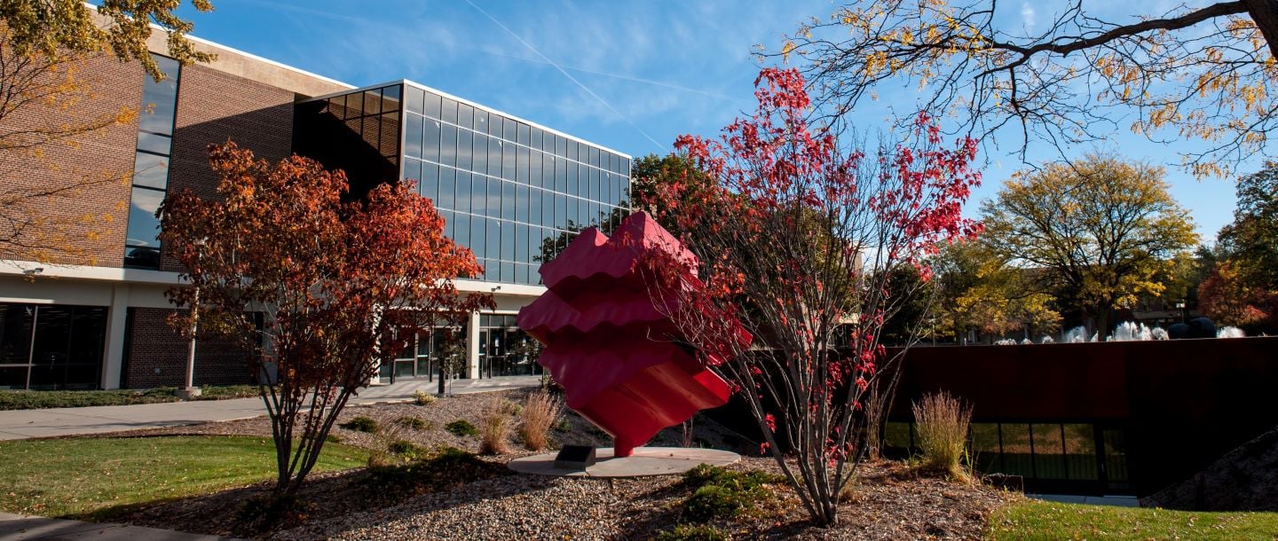 The outside area in front of Memorial Library on a sunny fall day with the red steel sculpture, titled Waves, in the background between two shrubs