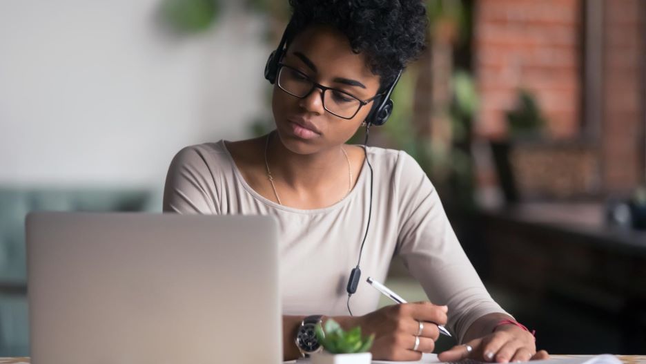 A female student studying at a table with a laptop taking notes and wearing headphones